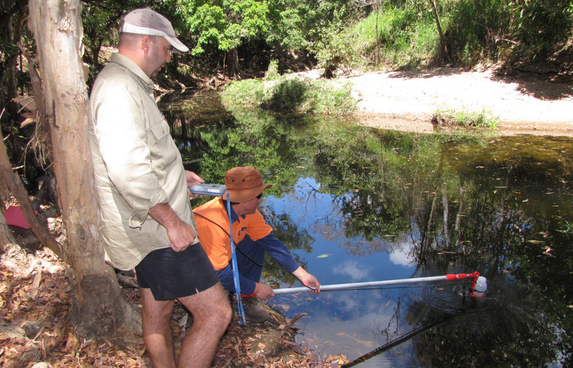 Two men are near a creek bed. The one standing wears a beige shirt and black shorts and holds a monitoring device. The other wears orange his-vis gear and is crouched down holding a stick into the middle of the water. 