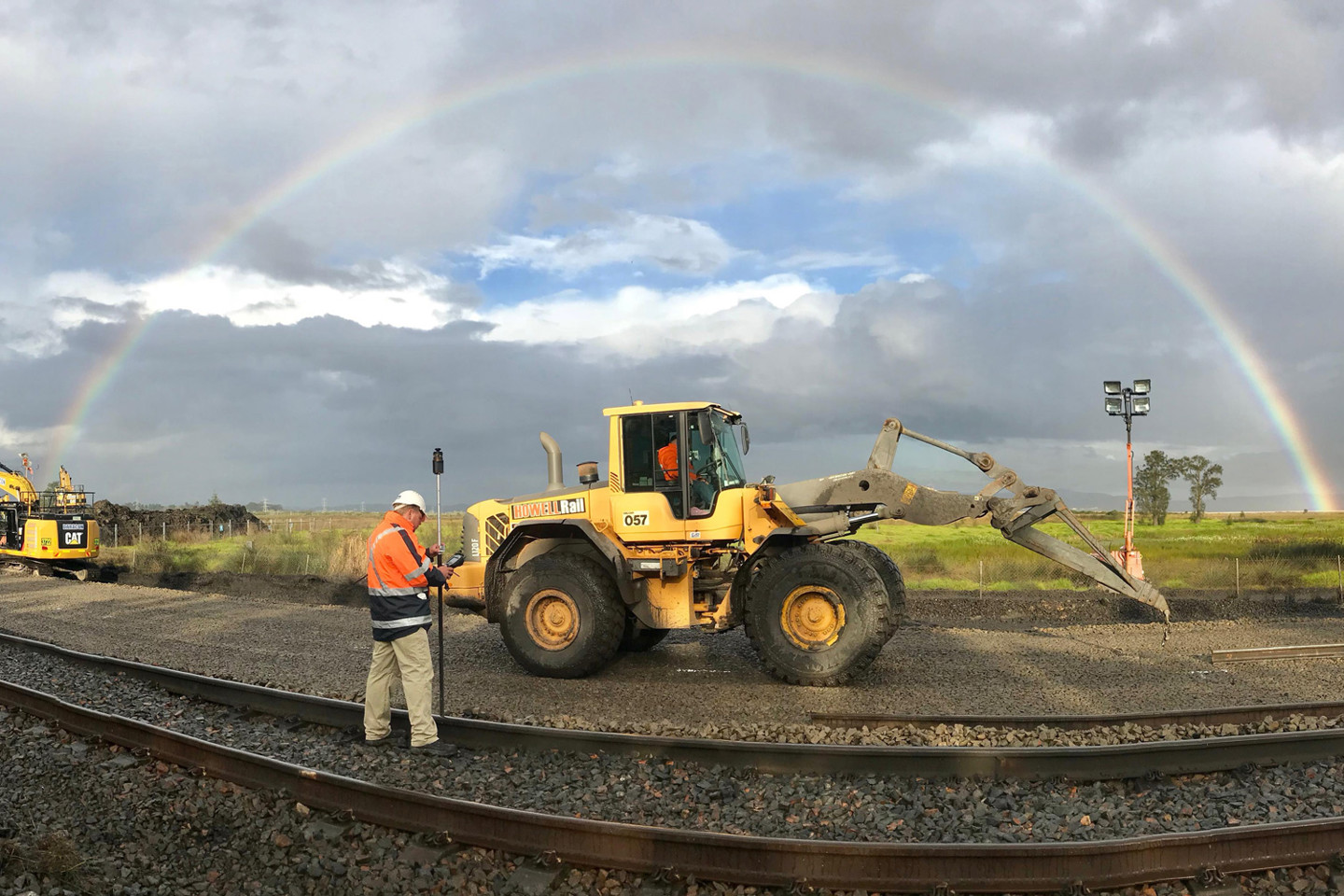 RPS surveyor stands in the middle of railway tracks with a digger in front of him and a large rainbow in the distance. 