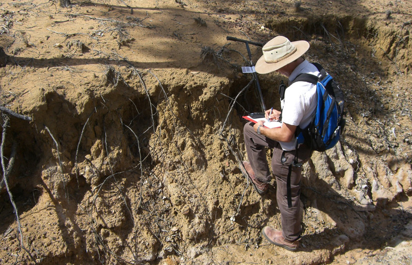 An RPS ecologist wearing hiking boots, cargo pants, and carrying a backpack stands in a mound of soil with a measuring device and a notepad taking notes. 